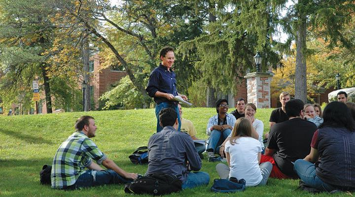 Teacher standing talking to a group of students sitting in the grass outside